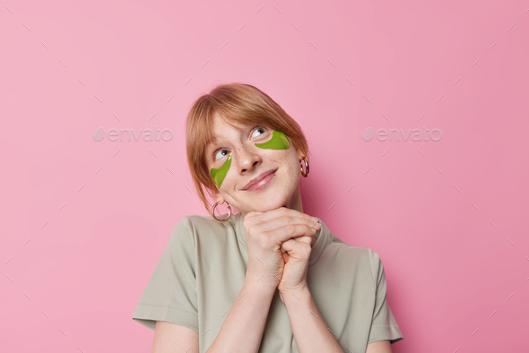 Horizontal shot of pretty dreamy woman keeps hands under chin looks above with satisfied expression