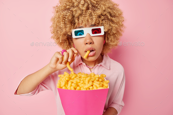 Photo of amazed woman holds breath as watches horror film in cinema eats delicious corn sticks from