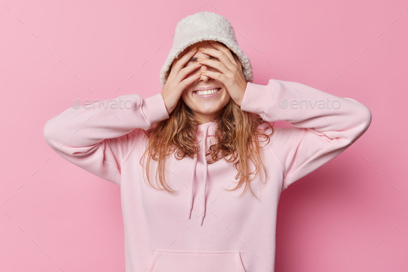 Happy delighted young woman hides face with hands smiles toothily wears hat and sweatshirt giggles p