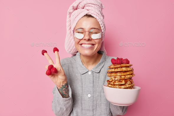 Positive young woman has fun going to eat yummy pancakes with raspberries and honey makes peace gest