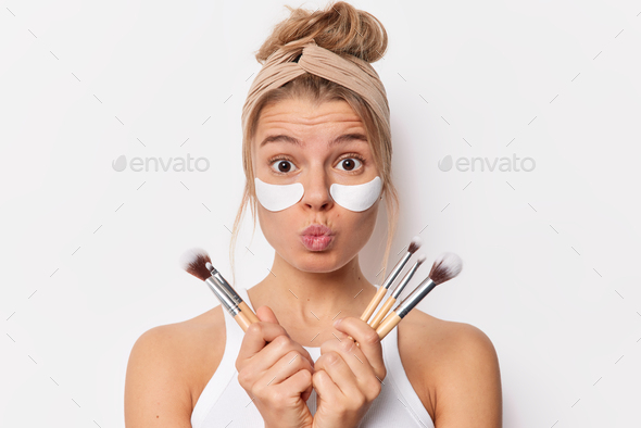 Surprised tender woman keeps lips folded holds cosmetic brushes applies beauty patches under eyes to
