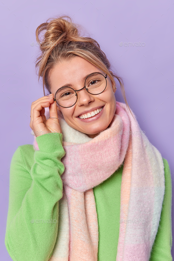Indoor shot of lovely young fair haired woman tilts head keeps hand on rim of spectacles smiles broa