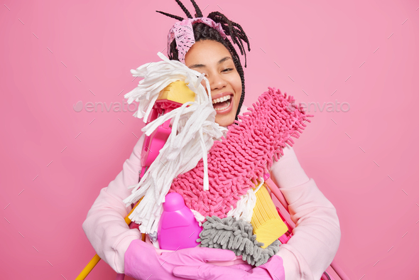 Positive woman embraces big heap of cleaning tools and detergents ready for tidying up house wears r