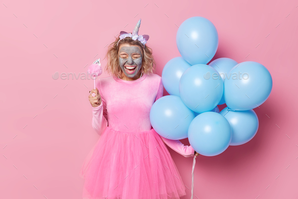 Happy young woman wears festive dress and unicorn headband applies clay mask for reducing fine lines