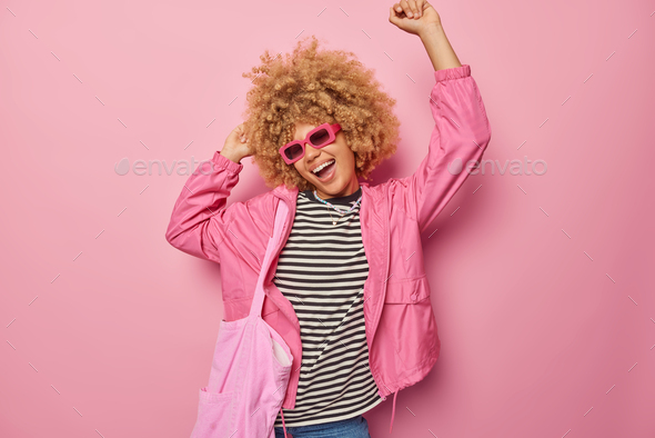 Cheerful woman with curly hair moves with rhythm of music keeps arms raised dances carefree dressed