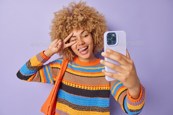 Beautiful positive woman makes peace gesture over eye sticks out tongue winks eye poses at smartphon