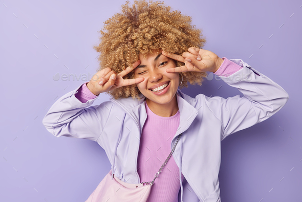 Pretty positive woman with curly bushy hair makes peace gesture with both hands smiles gladfully wea