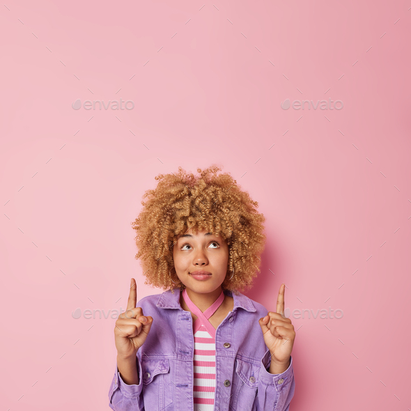 Serious young woman with curly hair points overhead with both index fingers advertises product dress