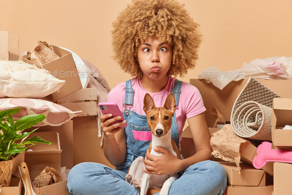 Funny European woman with curly hair sits on floor with dog and uses smartphone moves in new apartme