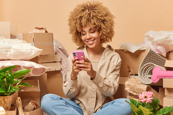 Young curly haired smiling woman uses smartphone scrolls newsfeed sits on floor with cardboard boxes
