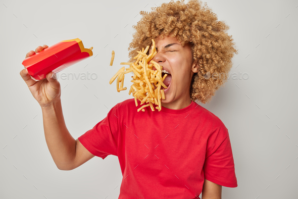 Cheat meal concept. Curly haired woman eats french fries containing much calories dressed in casual