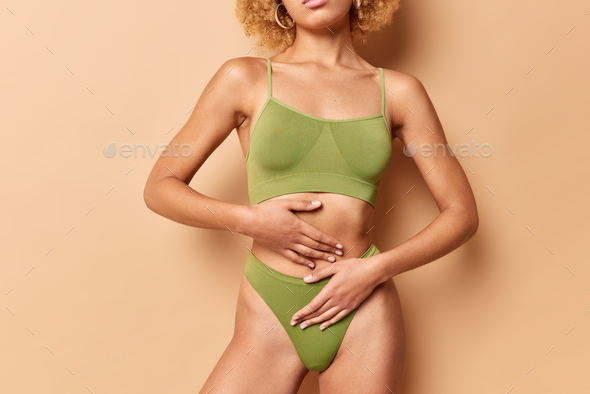 Cropped view of unrecognizable fit woman wears green top and panties keeps hands on flat belly poses