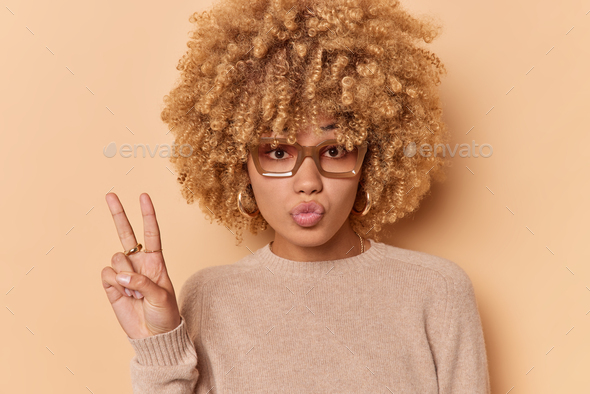 Lovely cheeky beautiful young woman with curly bushy hair makes peace sign keeps lips folded wears c