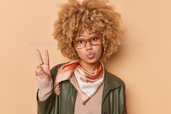 Photo of romantic young woman keeps lips folded blows mwah makes peace gesture dressed in fashionabl