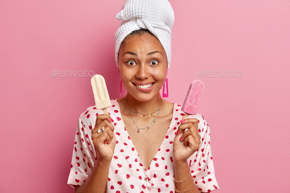 Horizontal shot of cheerful young woman holds two gelatos bites lips suggests friend to eat ice crea