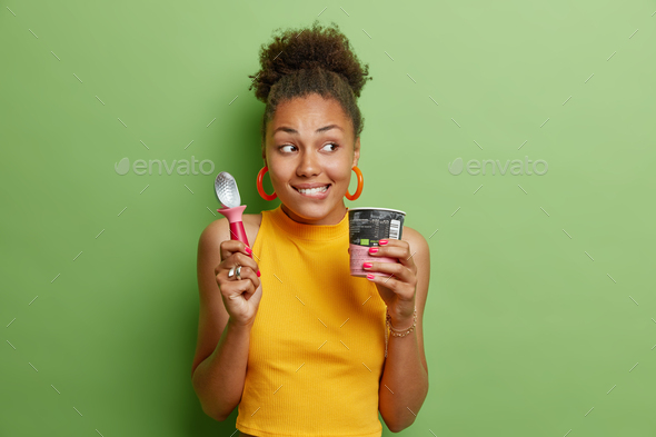 Photo of curly haired young woman enjoys eating cold ice cream with spoon during summer day bites li