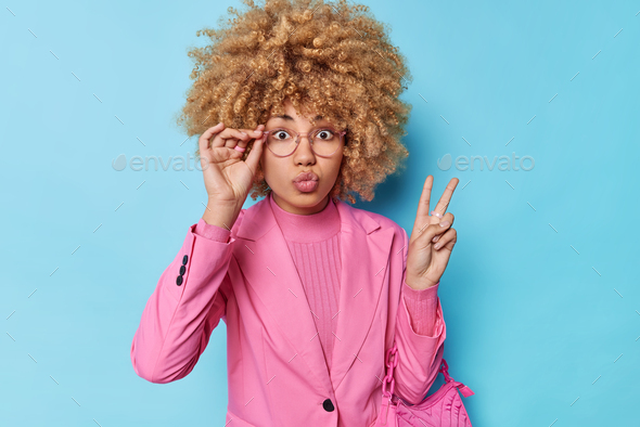Stay positive. Curly haired woman keeps lips folded makes peace gesture keeps fingers raised assures