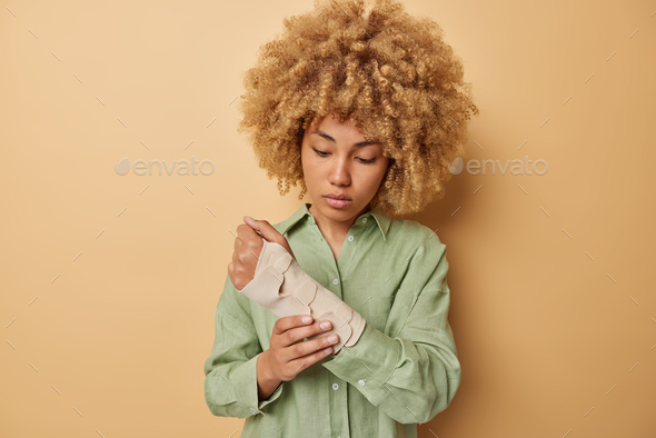 Serious curly haired woman applies elastic bandage on wounded arm got injured or trauma wears green