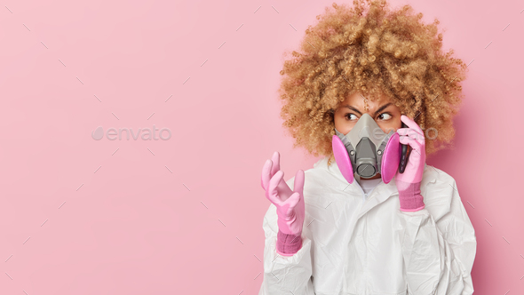 Angry young woman wears gas mask and bio hazard suit makes telephone call gestures with annoyance wo