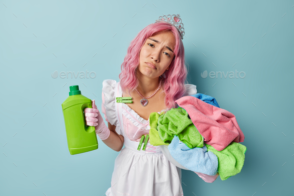 Exhausted maid feels tired of housework poses with dirty laundry and chemical softener detergent loo