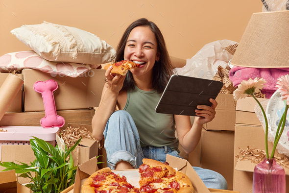 Happy young Asian woman eats pizza holds modern tablet surfs internet seatrches ideas for house deco
