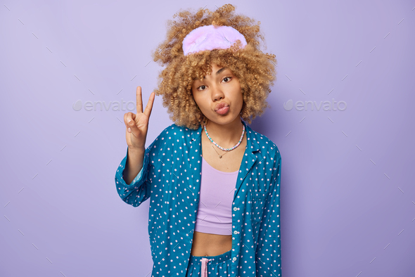 Indoor shot of curly haired woman wears sleepmask and comfortable pajama keeps lips folded gestures