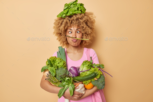Indoor shot of funny woman makes grimace poseswith spring harvest embraces bunch of different freshl