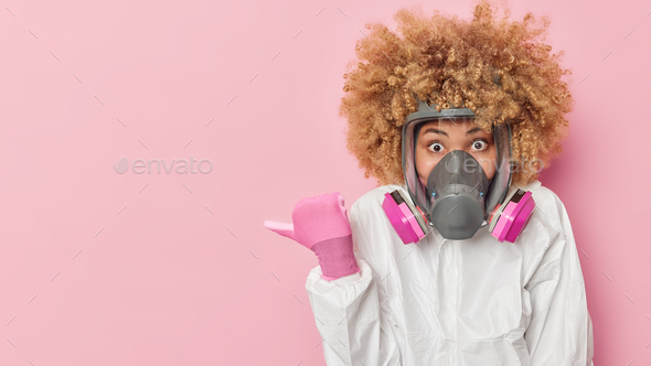 Impressed curly woman scientist wears coronavirus protection suit and mask points thumb away on mock