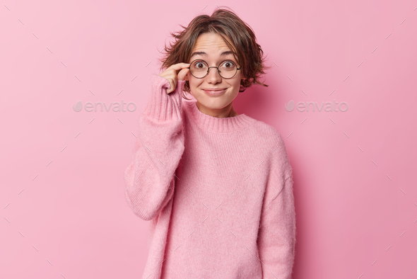Positive surprised woman keeps hand on rim of spectacles smiles pleasantly reacts on good news wears