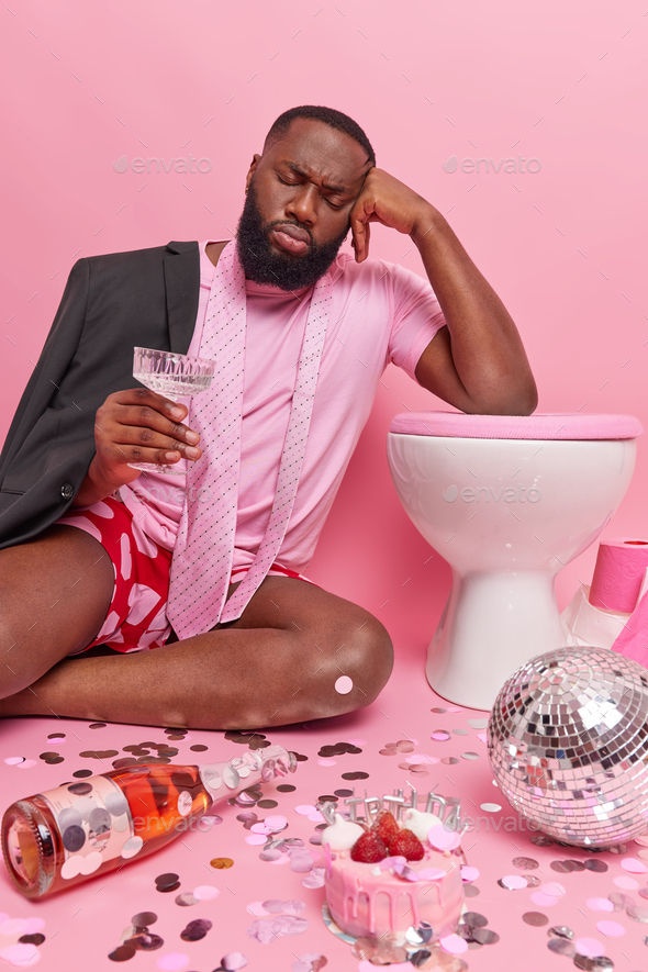 Sleepy dark skinned man tired after party holds glass of cocktail leans on toilet bowl has mess in r