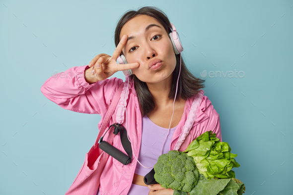 Lovely brunette Asian woman makes peace gesture keeps lips folded carries green vegetables being veg