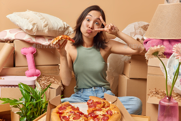 Young Asian woman keeps lips rounded makes peace gesture over eye eats tasty pizza sits crossed legs