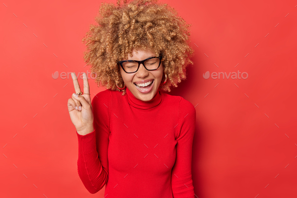 Overjoyed curly haired woman laughs happily makes peace gesture enjoys life keeps eyes closed being