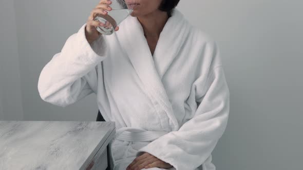 Thirsty smiling 35s woman in white bathrobe drinking water