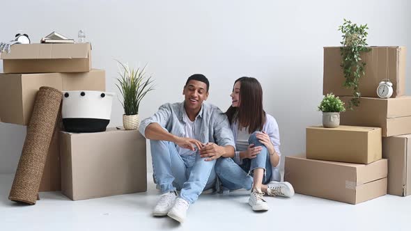 Happy Young Multiracial Couple Caucasian Woman and Hispanic Guy are Sitting on the Floor Between alt