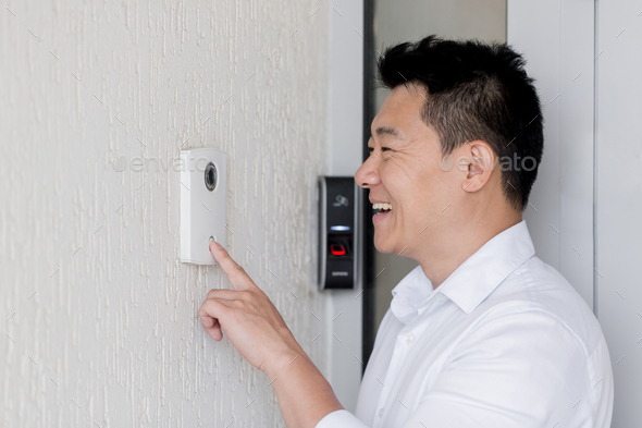 A young Asian man rings an electronic doorbell on the wall of a house with his finger. He opens the