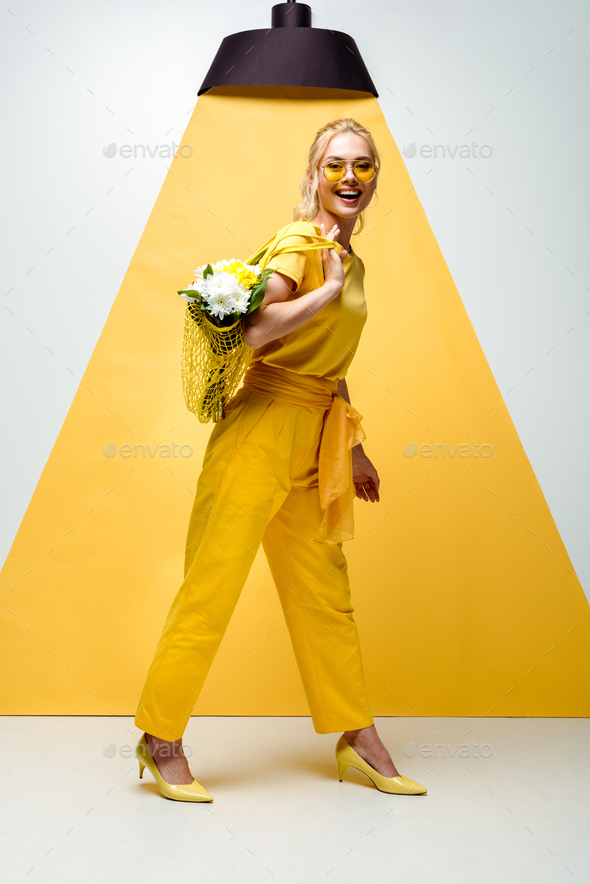 cheerful blonde woman holding string bag with flowers while posing on white and yellow