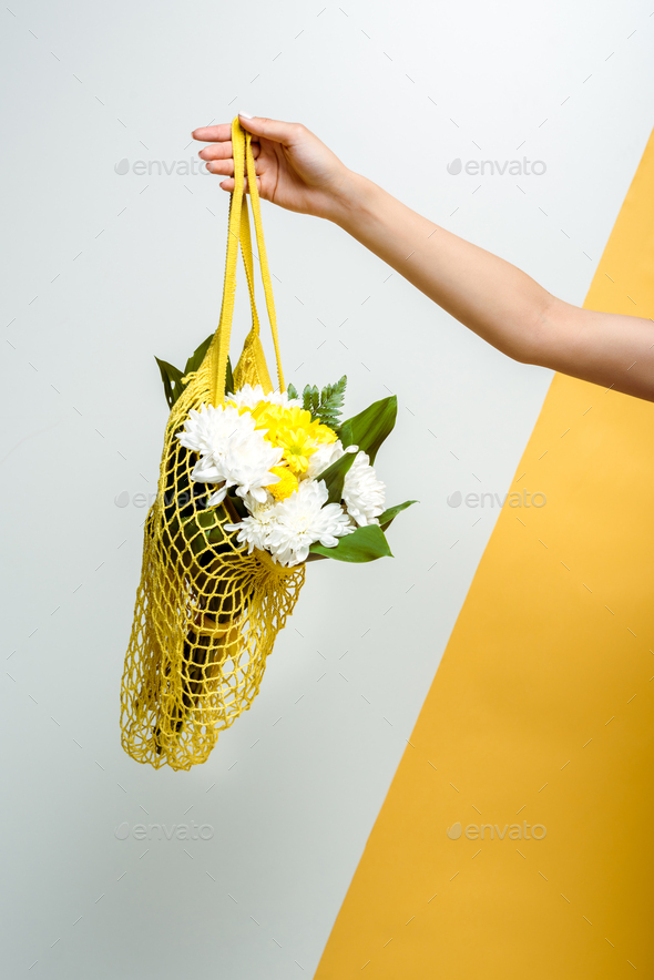 cropped view of woman holding string bag with flowers on white and yellow
