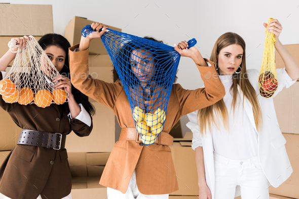 african american women covering faces while holding string bags with fruits near blonde girl on