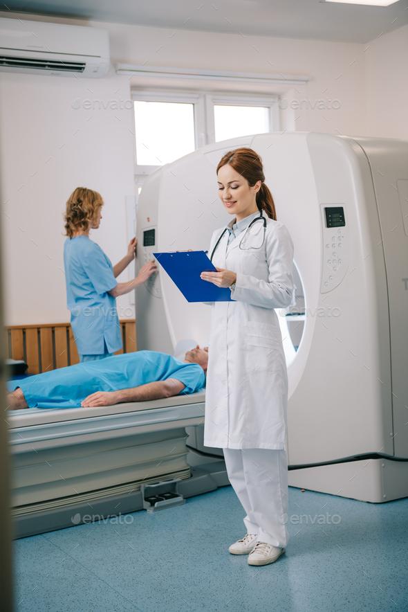 smiling radiographer writing on clipboard while assistant standing near ...