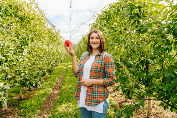 A woman farmer with an apple in her hands stands between even rows of fruit trees smiling.