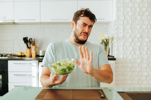 Young man is fed up healthy food in the form of greens vegetables grimaces in front of this food.