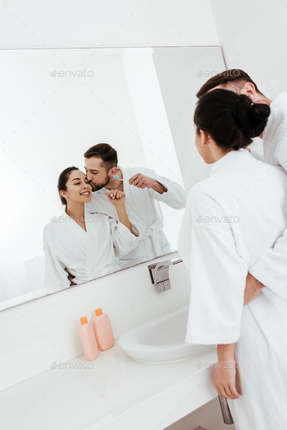 bearded man kissing cheek of cheerful woman looking at mirror in bathroom