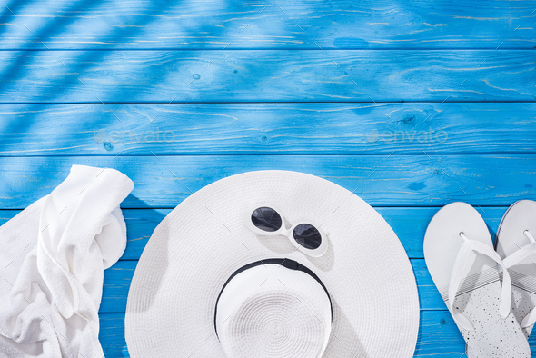 top view of crumpled towel, white glasses, flip flops and floppy hat on blue wooden background with