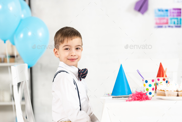 adorable boy sitting at party table and looking at camera during birthday celebration