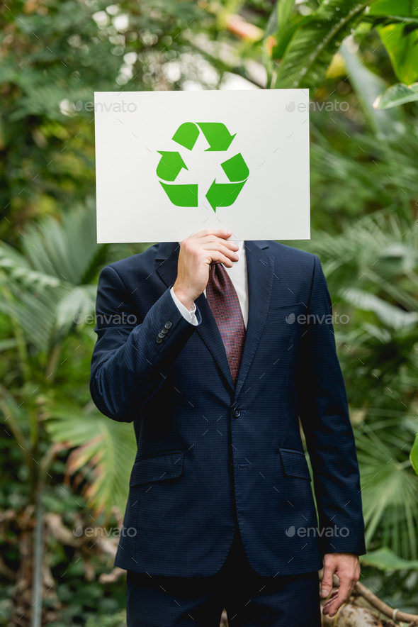 businessman in suit holding card with green recycling sign in front of face in greenhouse