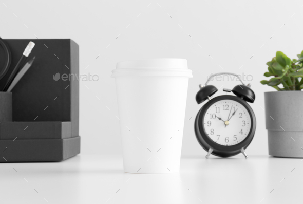 Coffee paper cup mockup with a clock, workspace accessories and a succulent plant on a white table.