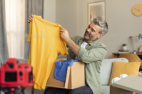 Cheerful elderly caucasian male blogger shoots vlog on camera with empty screen opens parcel shows