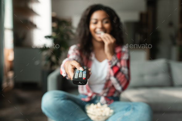 Happy millennial black lady with remote control change channel on tv, watch movie, eat popcorn
