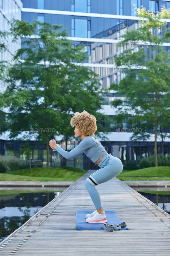 Outdoor shot of sporty woman dressed in blue tracksuit does squats with resistance band has workout